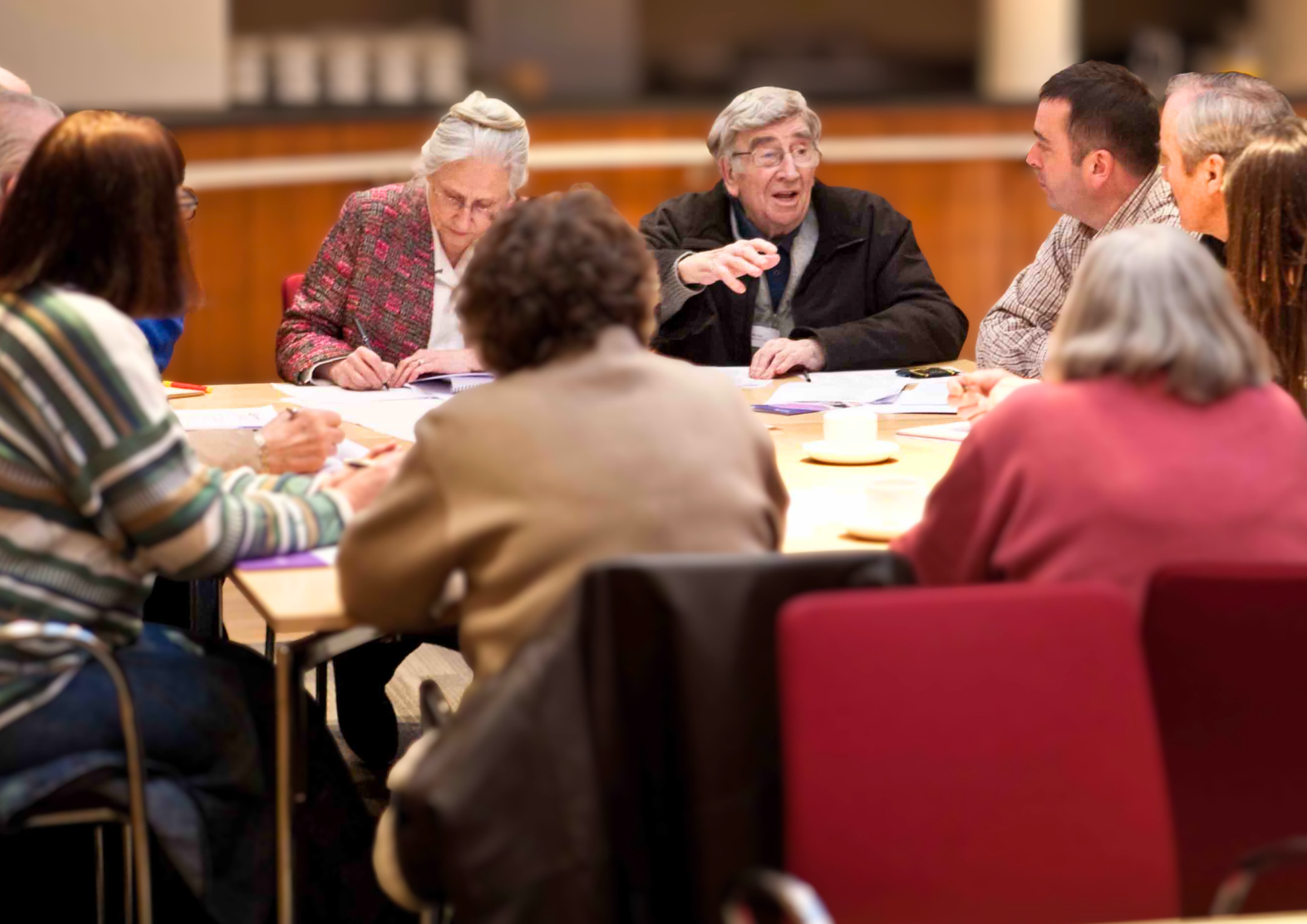 A group of adults seated around a large table in a training session. A man at the centre gestures as he speaks, while others listen and take notes.