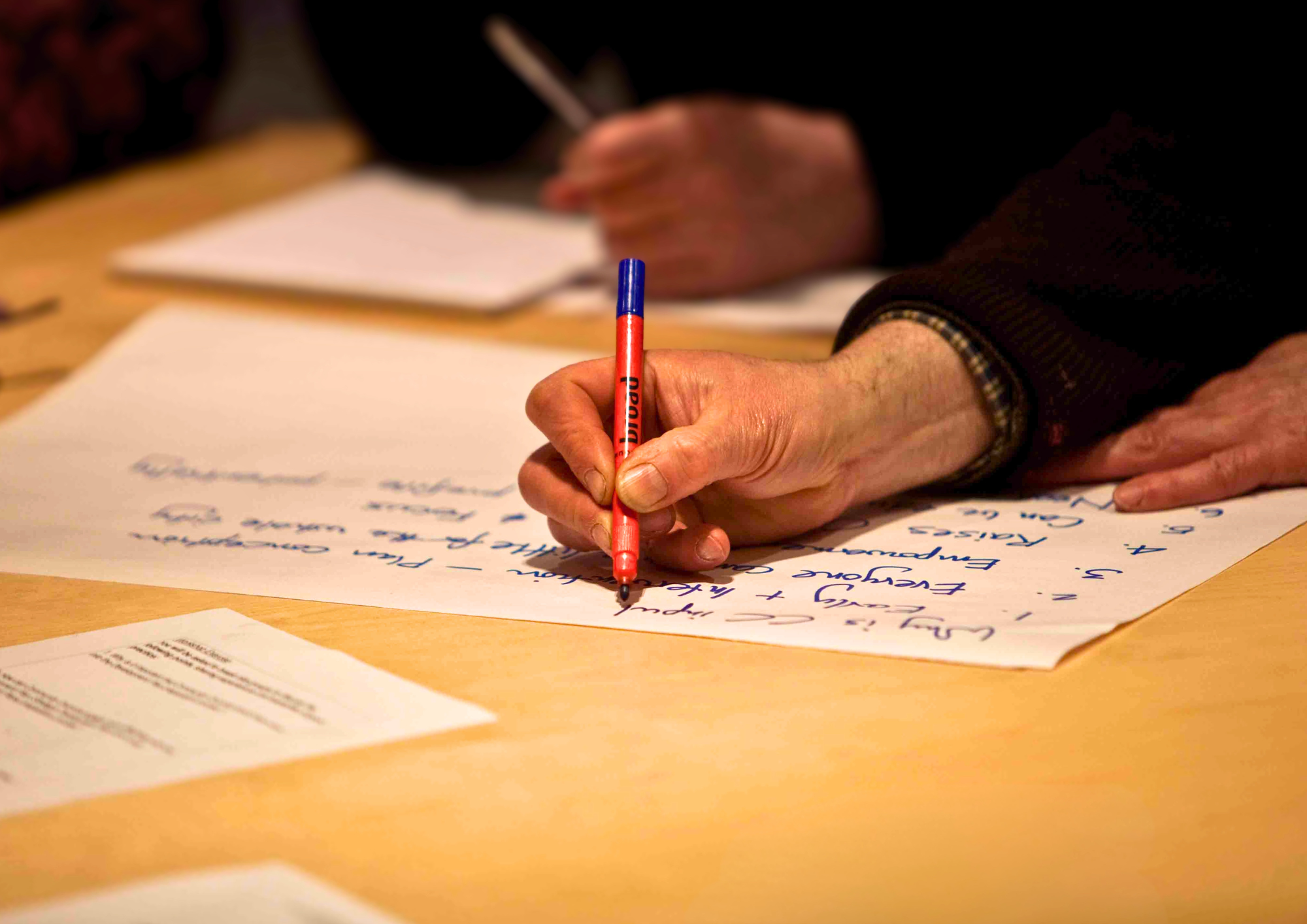 A close-up of a hand holding a red and blue marker pen, writing notes in blue ink on a sheet of white paper.